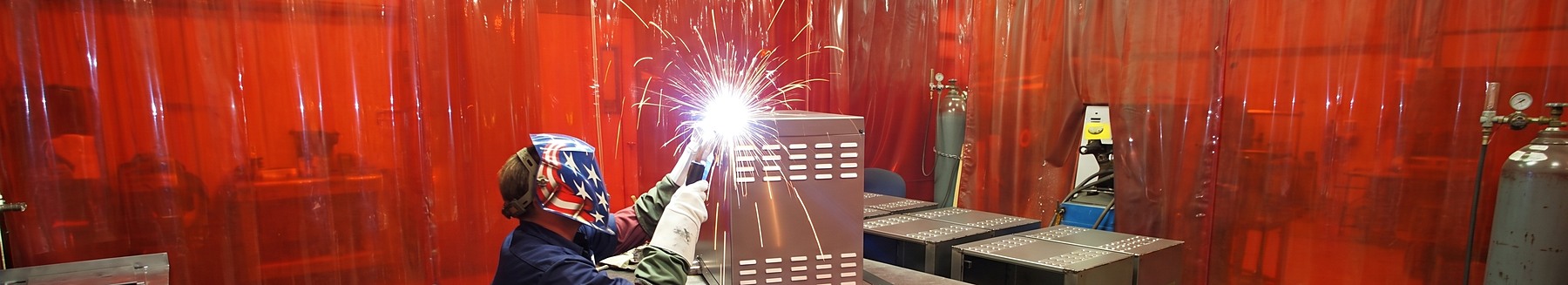 Atlas welder running a bead on a stainless enclosure, with finished enclosures staged behind